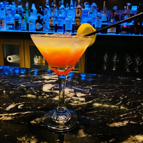A vibrant orange and yellow cocktail with a lemon slice and black straws on a dark marble bar.