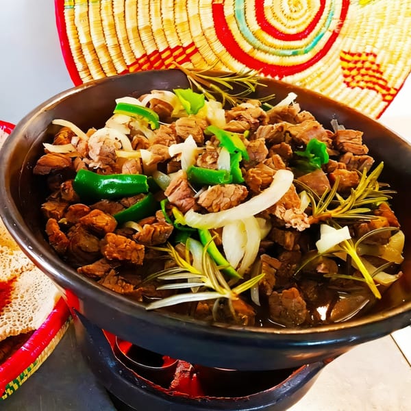 Ethiopian tibs, a dish of cubed meat with onions, green peppers, and rosemary, in a dark pot.